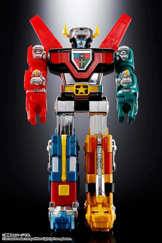 A multicolored robot toy figure standing against a dark studio background. The figure has a black torso with red wings, a red right arm, a teal left arm, blue and yellow legs, and a yellow chest emblem with a red cross.