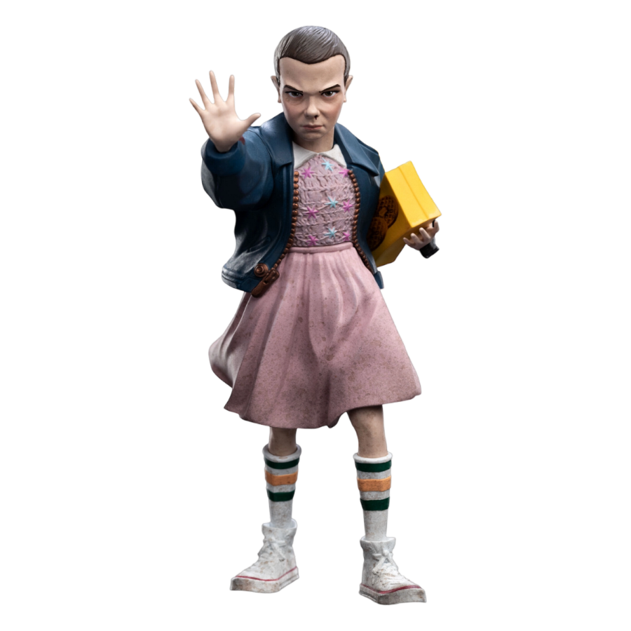 Action figure of Eleven from Stranger Thingsin a pink dress and blue jacket holding a yellow box on a white background. Stranger Things Eleven Mini Epics Vinyl Figure.